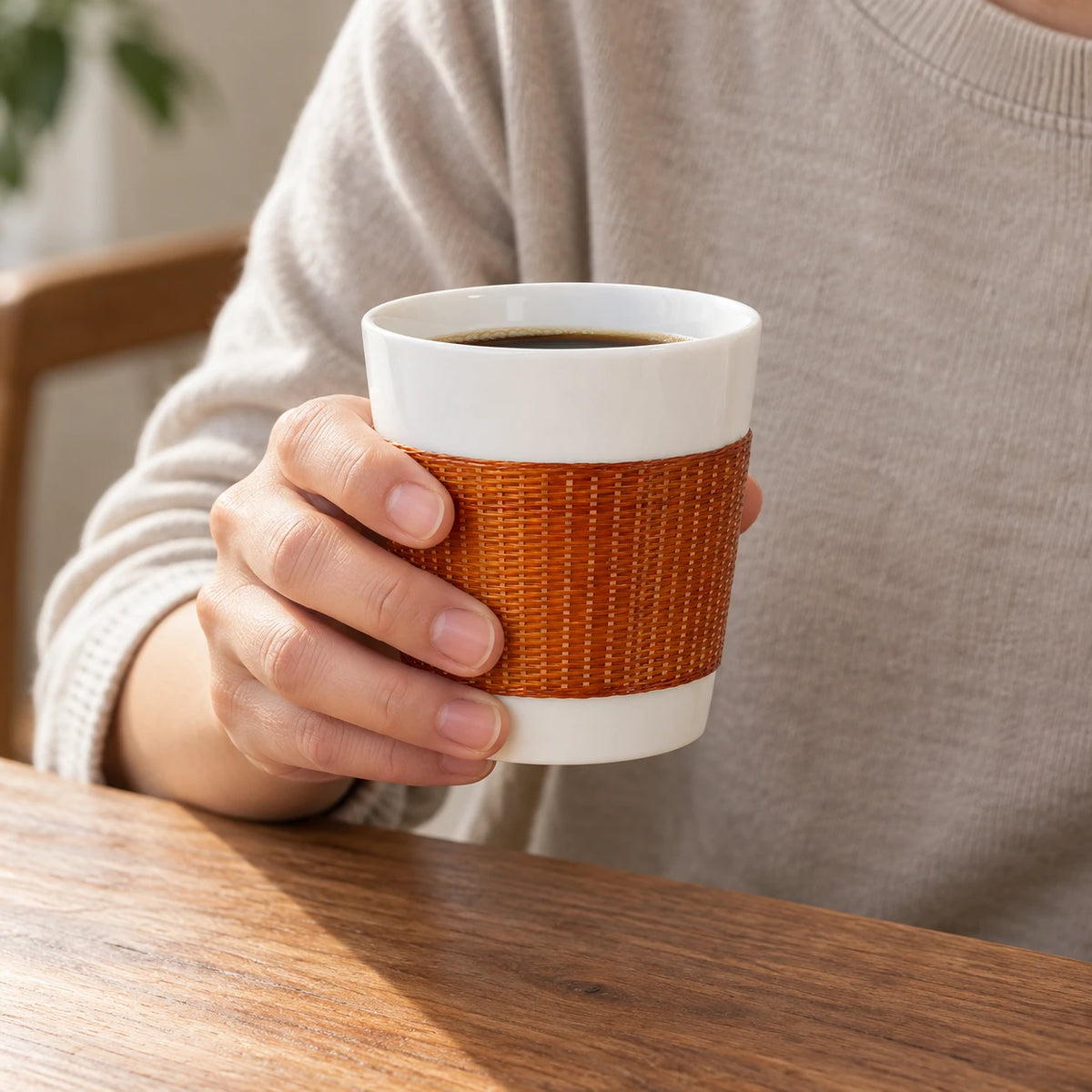 A person holding the YUNXI Daily Cup in Baked at a wooden table, showing its handwoven bamboo exterior, white porcelain rim and coffee inside.