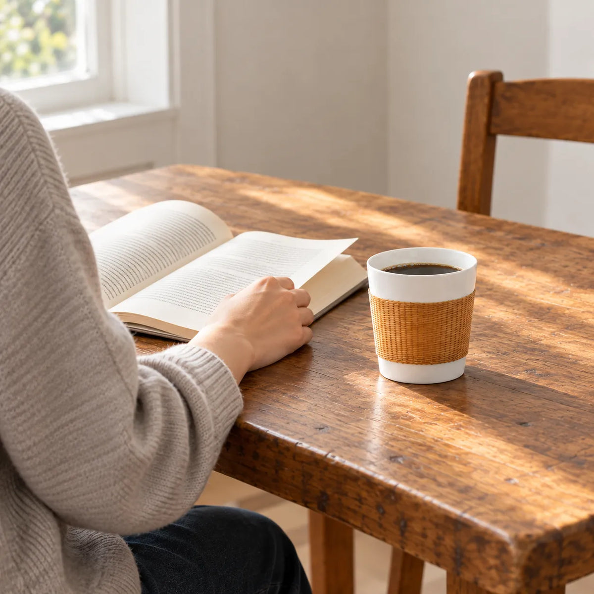 Natural YUNXI Daily Cup with handwoven bamboo and white porcelain, filled with coffee beside an open book as a person reads at a wooden table.